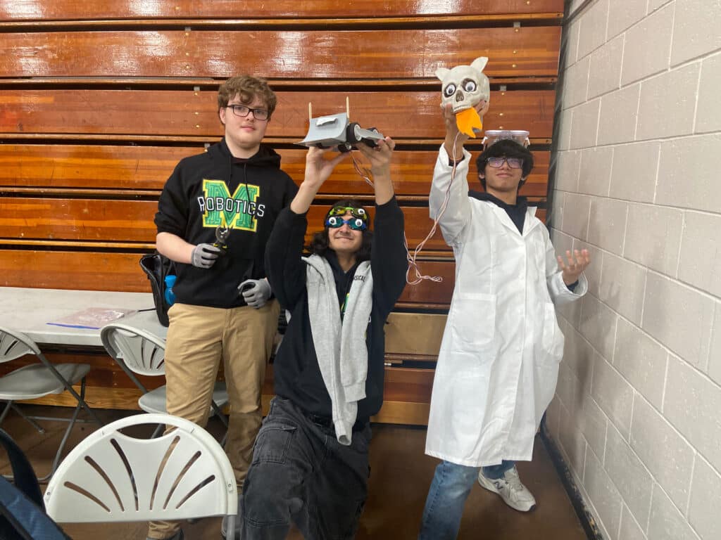 Three high school students hold their robot up to the camera. This robot features a skull shaped controller!
