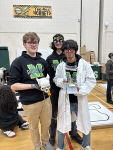 Three high school students hold their robot up to the camera. This robot features a skull shaped controller!